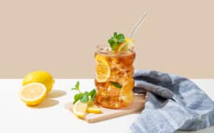 Iced tea with lemon, ice and mint in a glass on beige and white background with shadow and citrus fruits. Trendy concept of refreshing drink or lemonade, menu advertisement mockup. Front view.